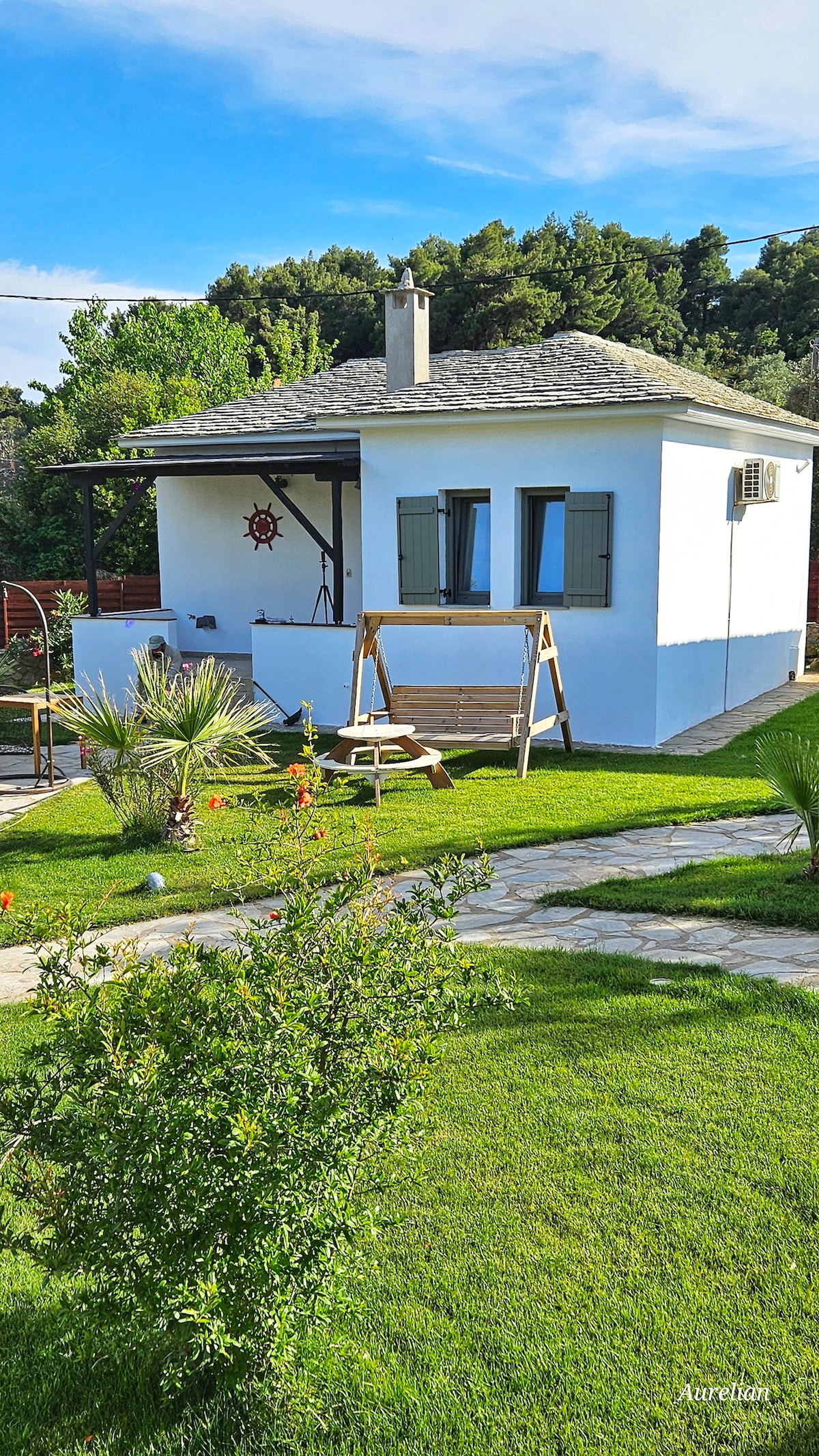 A charming white cottage with green shutters is situated amidst well-maintained gardens. A wooden swing set is positioned on the lawn, surrounded by lush greenery and colorful flowers. The pathway leading to the entrance is lined with natural stones, enhancing the inviting outdoor space.
