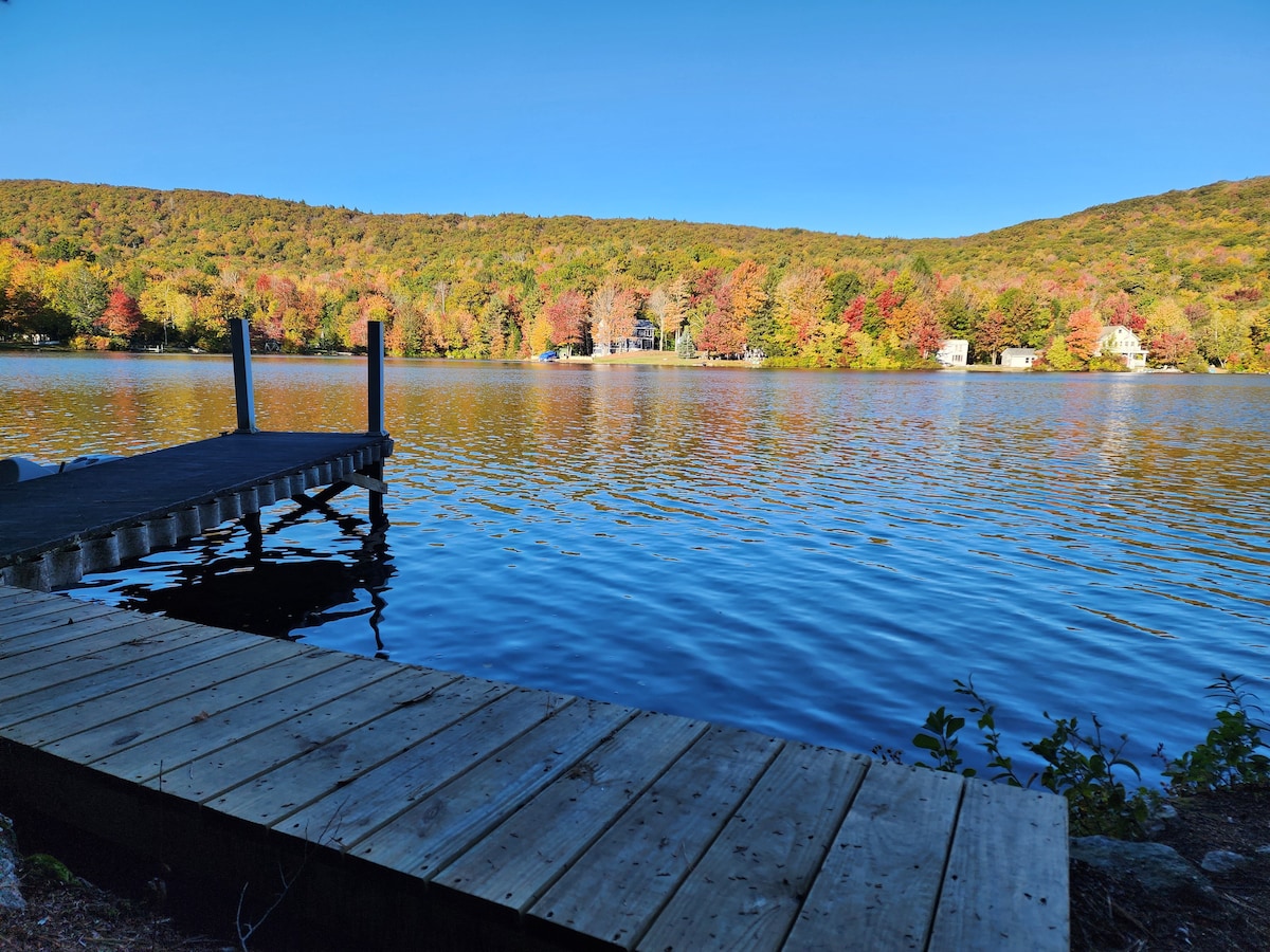 A wooden dock extends over calm waters, framed by vibrant autumn foliage. The reflection of trees in the water enhances the natural setting, while gentle ripples create a serene atmosphere. The distant shoreline reveals additional residences amid the colorful landscape.