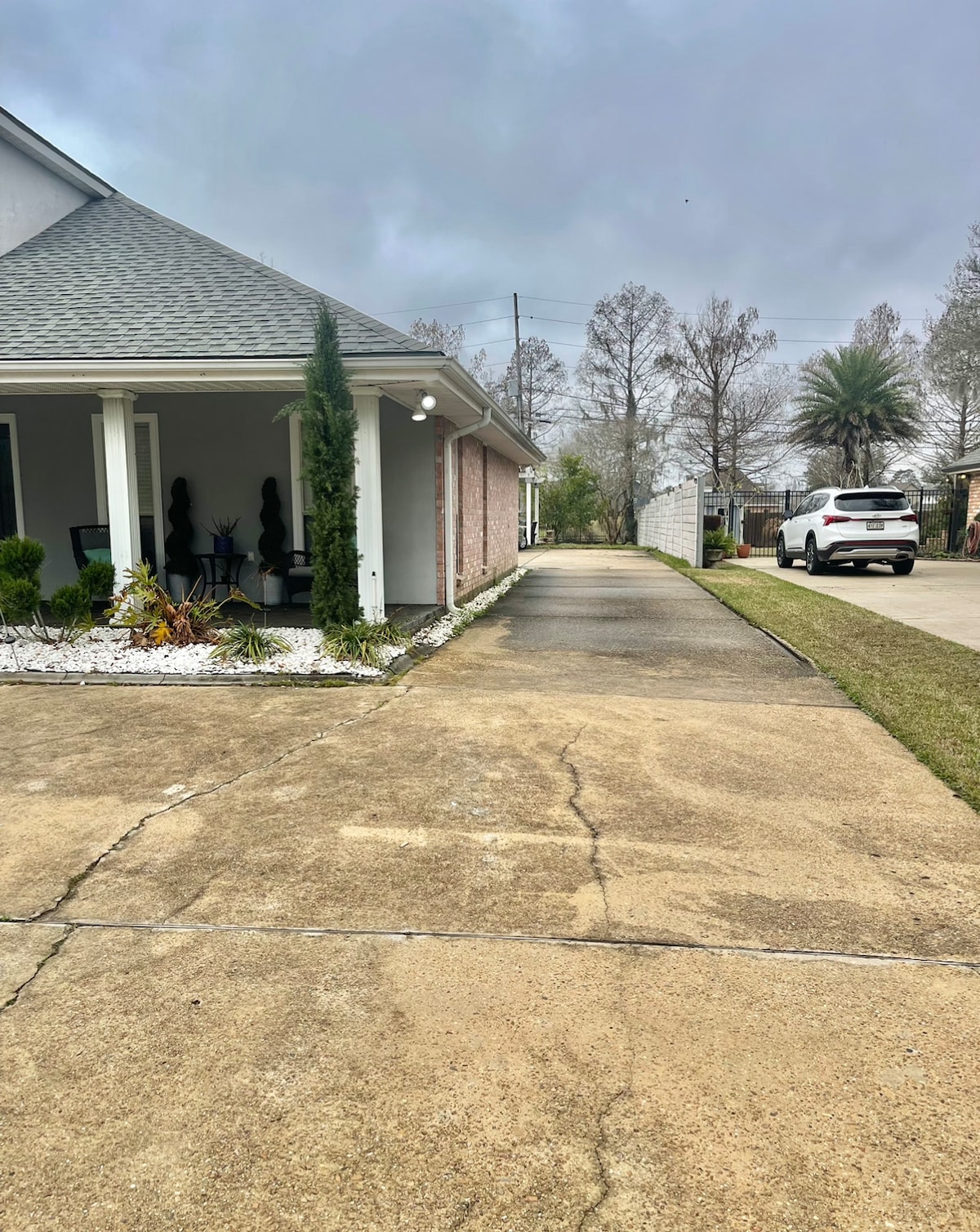 A driveway leads to the parking area, surrounded by neatly manicured shrubs and decorative stones. The side of the house is visible, featuring a light brick exterior and a covered porch with patio chairs. A vehicle is parked in the driveway.