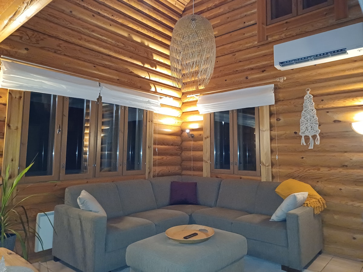 A cozy living area features a large, L-shaped sofa with a neutral colour scheme, complemented by a soft throw blanket. Natural wood walls and framed windows allow for abundant light, while a woven pendant light adds warmth to the space. A low coffee table completes the inviting setup.