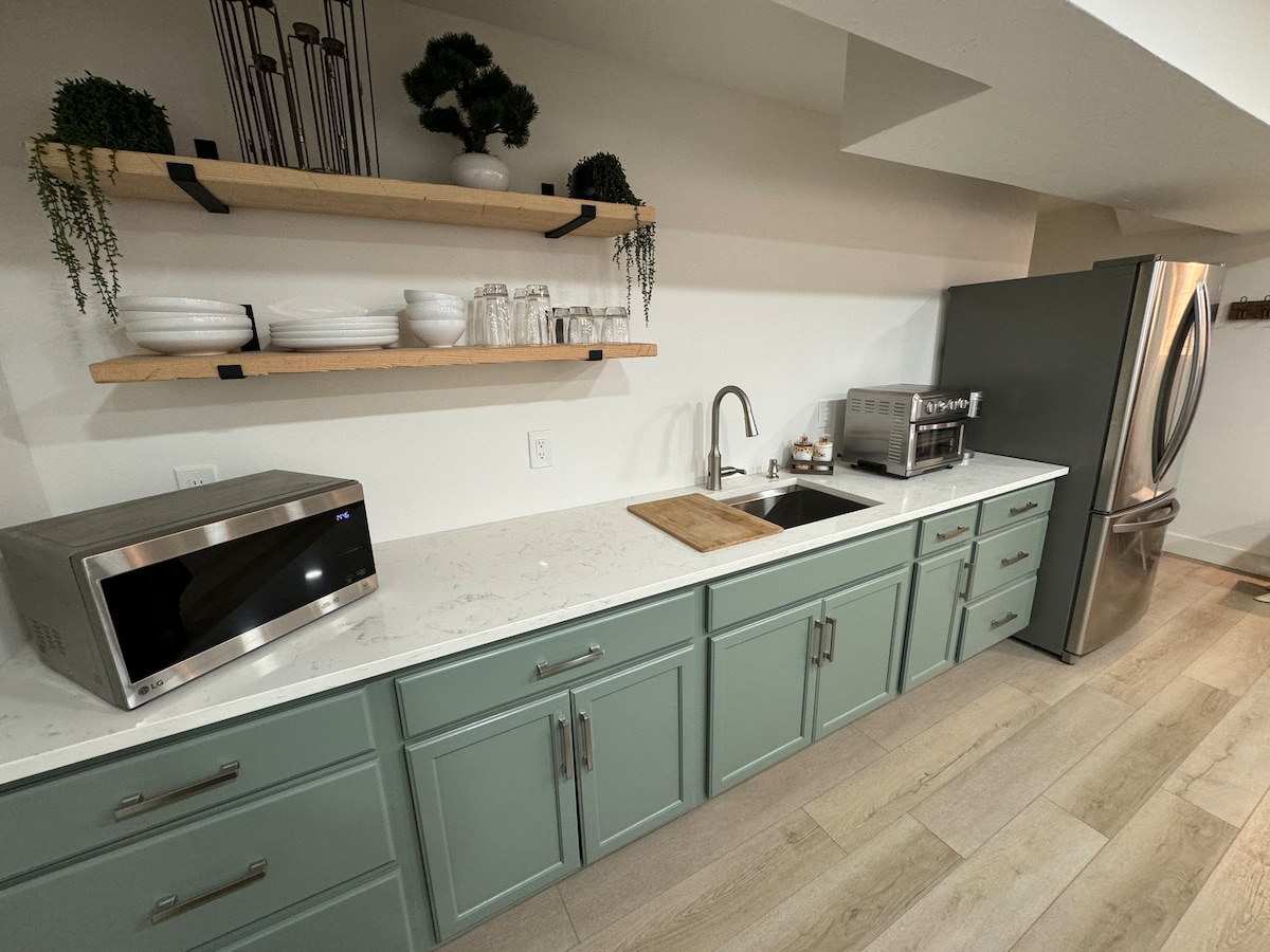 A well-appointed kitchen area features soft green cabinetry with a white countertop. Open shelving displays dishware and glasses. Appliances include a stainless steel refrigerator, microwave, and a sink with a modern faucet. Natural light highlights the clean, contemporary design.