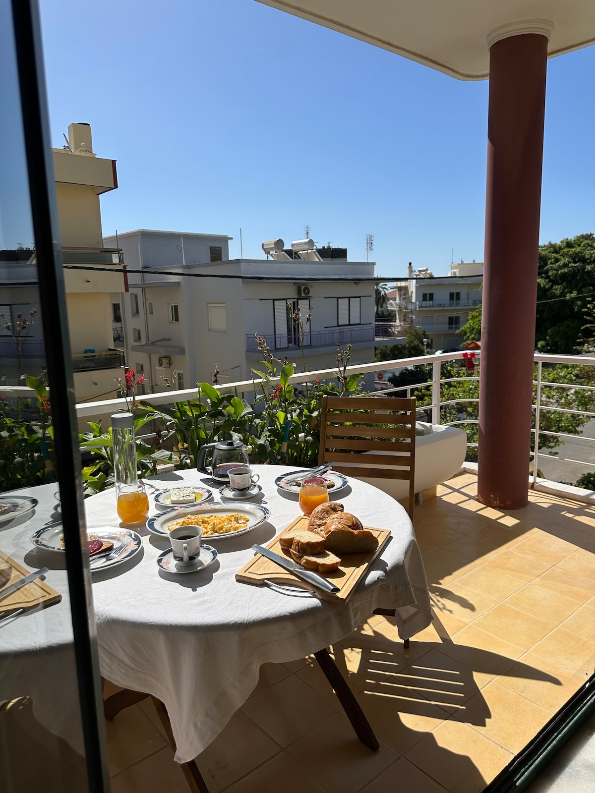 A balcony is set for breakfast with a round table displaying a selection of dishes including pastries and beverages. Nearby, a wooden chair complements the setting. Green plants provide a natural touch, and the bright sky is visible in the background.