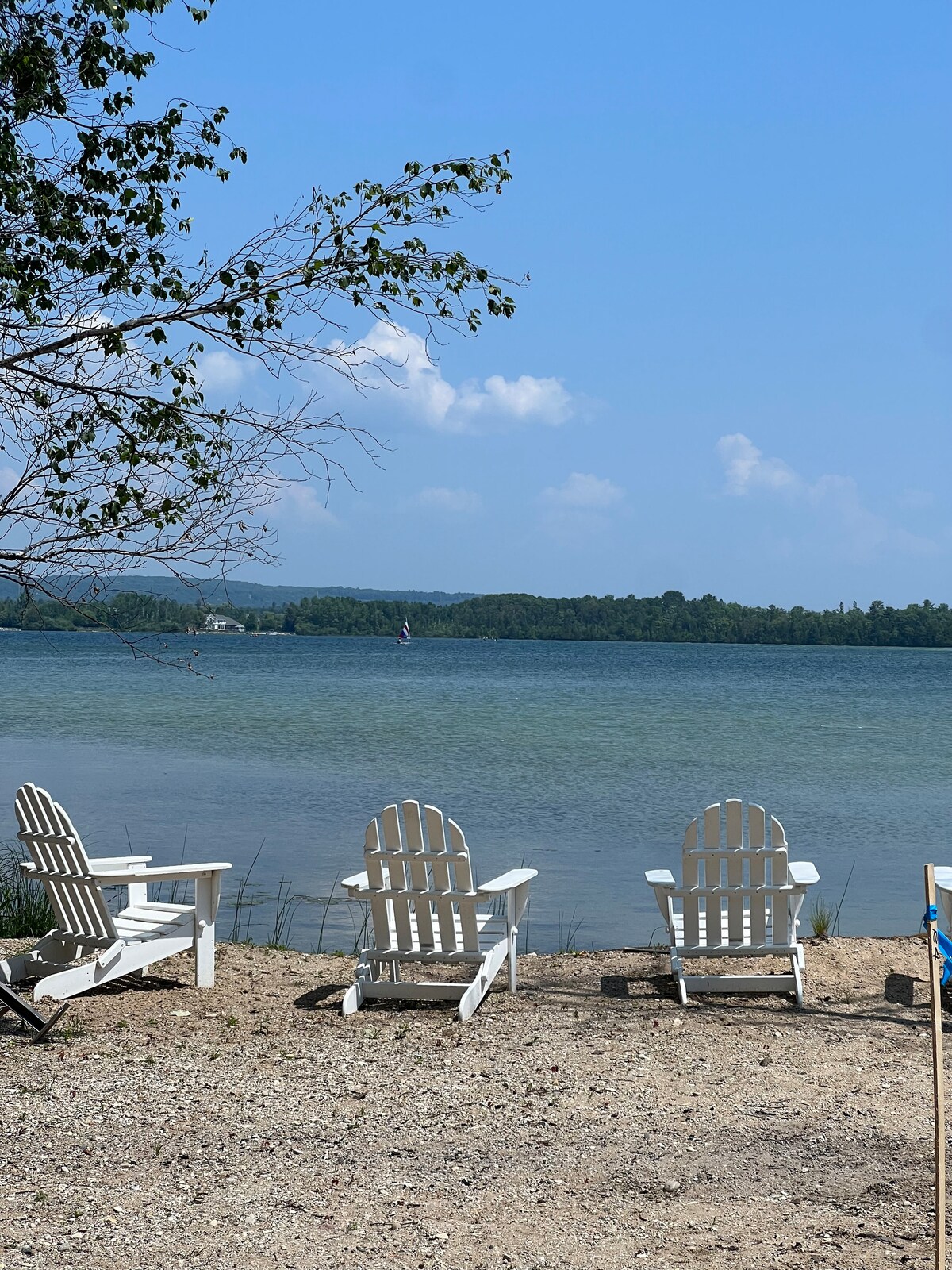 Adirondack chairs are positioned on a sandy shore by Round Lake, facing calm waters. The shoreline is framed by gentle hills in the background, with a clear blue sky overhead. The scene conveys a peaceful lakeside atmosphere, ideal for relaxation.