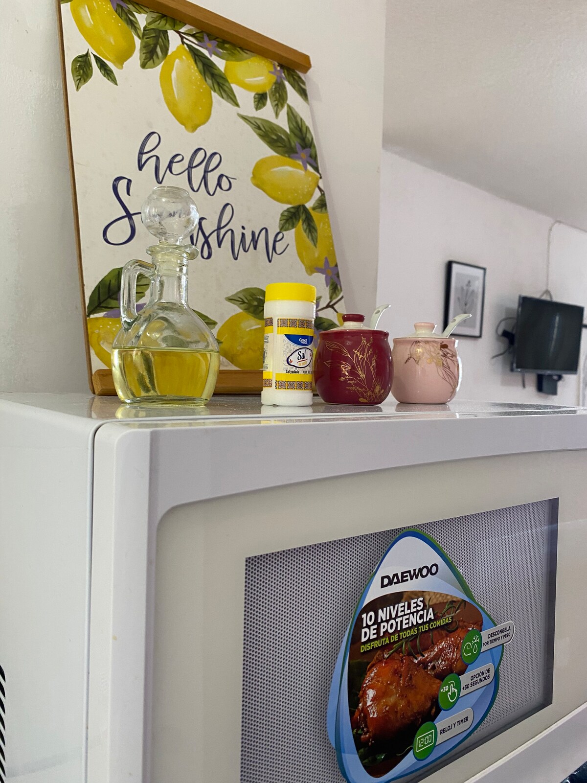 A kitchen counter features a white microwave with clear display graphics. On top, a decorative sign with the text 'hello Sunshine' is accompanied by a bottle of oil, a container of seasoning, and two small jars, each with a spoon for serving.