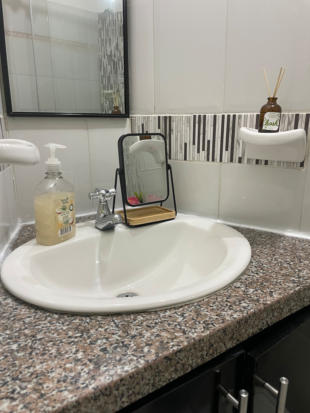 A clean bathroom sink area features a circular basin set into a speckled granite countertop. A small mirror is positioned on a wooden stand beside a bottle of hand soap, while a decorative item sits on the wall-mounted shelf nearby.