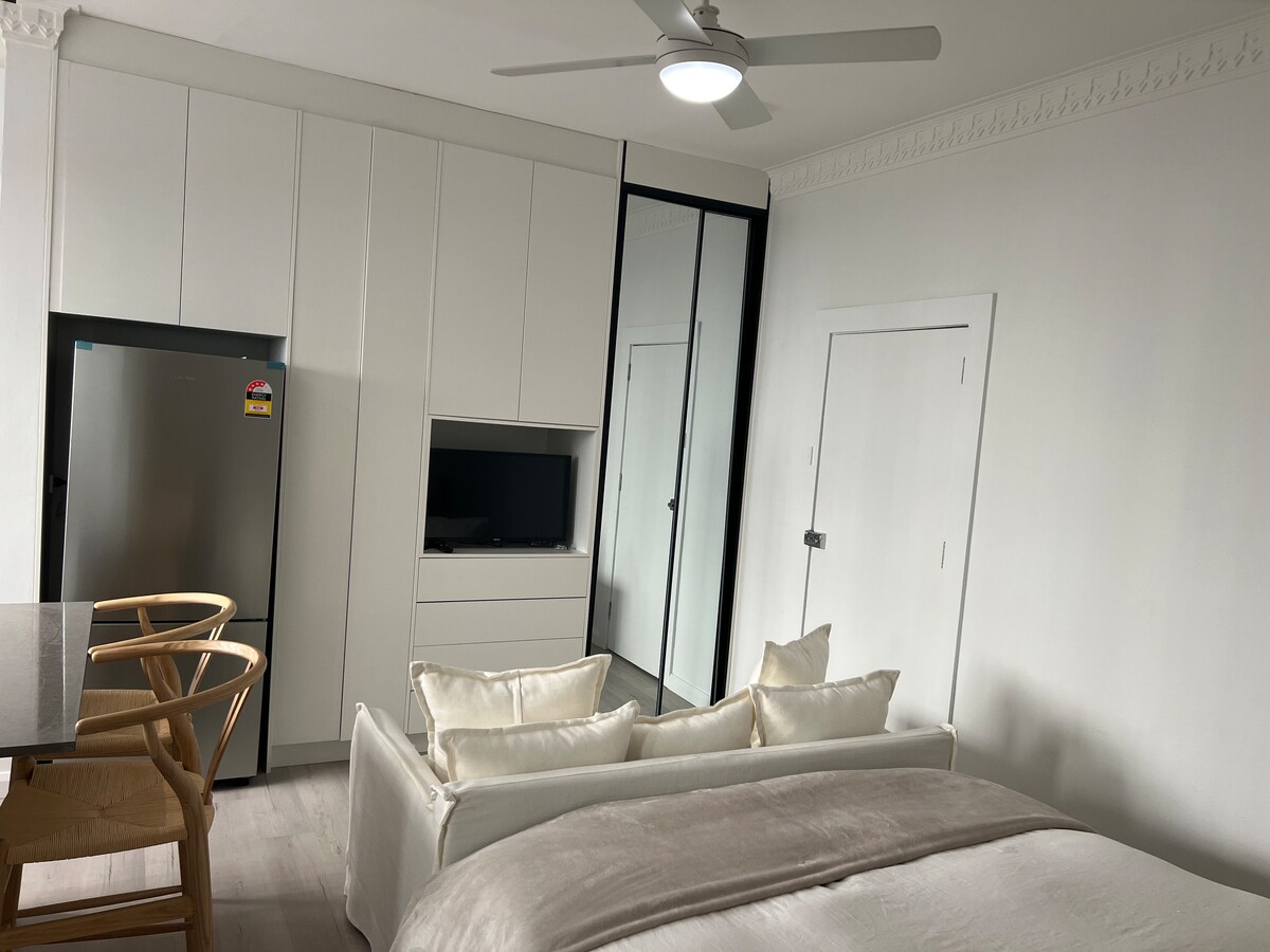An open studio layout is observed, featuring a spacious bed with neutral bedding. A sleek kitchenette is positioned alongside a mirrored closet, while a small dining area with two wooden chairs is present. Bright lighting is provided by a ceiling fan and wall fixtures.