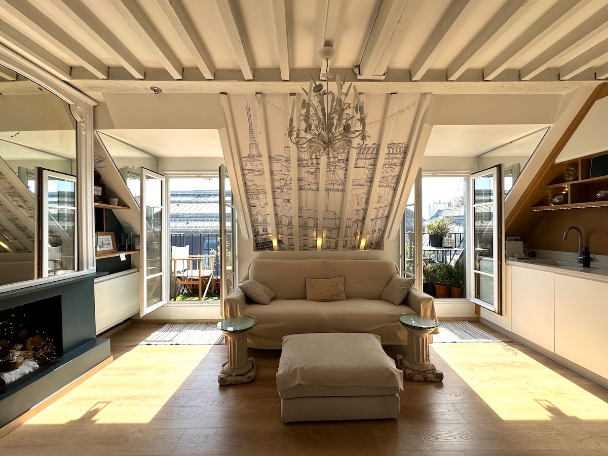 The living area features a light-colored sofa centered beneath a decorative chandelier. Large glass doors provide access to the balconies, while a stylish mural depicting Parisian architecture adds charm to the room. A pouf is positioned in front of the sofa, enhancing the comfortable setting.