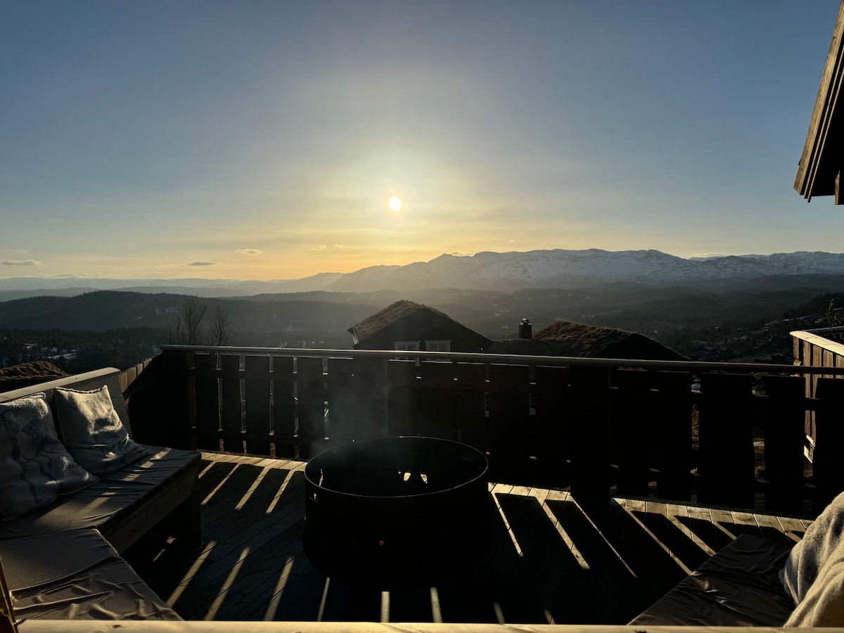 A spacious outdoor area features comfortable seating, with a view of a sunset illuminating the expansive landscape. The horizon showcases rolling hills, with snow-capped peaks in the distance. A fire pit is positioned in the center, inviting relaxation under the open sky.