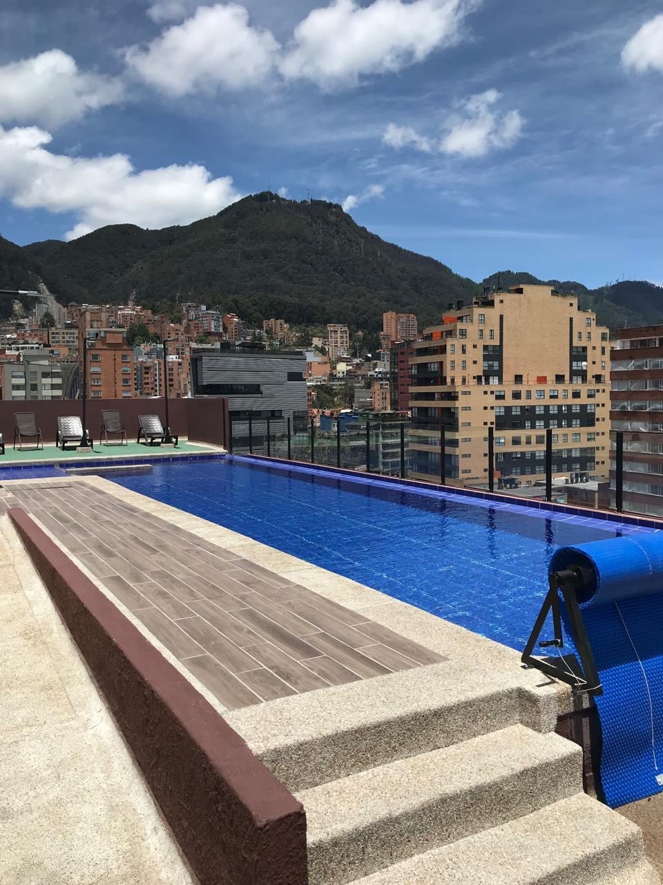 A rooftop swimming pool with a sleek, modern design overlooks a mountainous landscape and a city skyline. Sun loungers are placed along the pool's edge, and steps lead down into the water. Clear blue skies provide a bright backdrop.