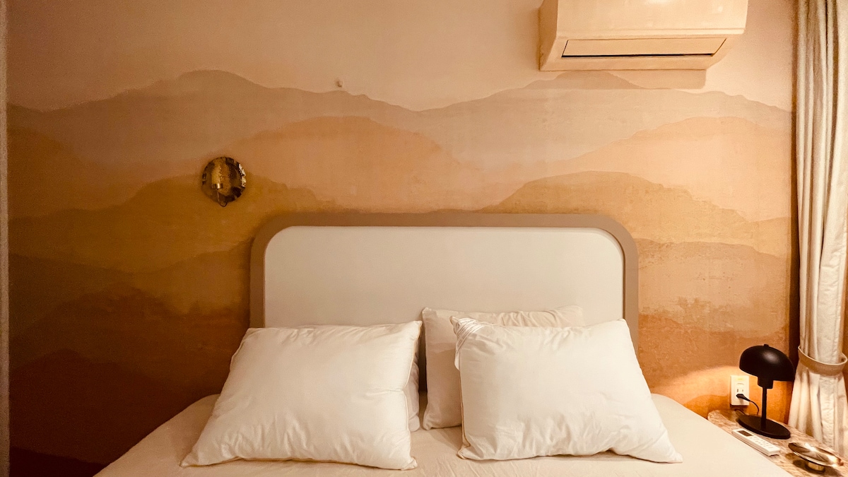 A tranquil sleeping area features a soft bed with plush white pillows against a backdrop of elegantly painted walls in warm earth tones, reminiscent of distant hills. Ambient lighting comes from a stylish lamp positioned next to the bed, enhancing the serene setting.