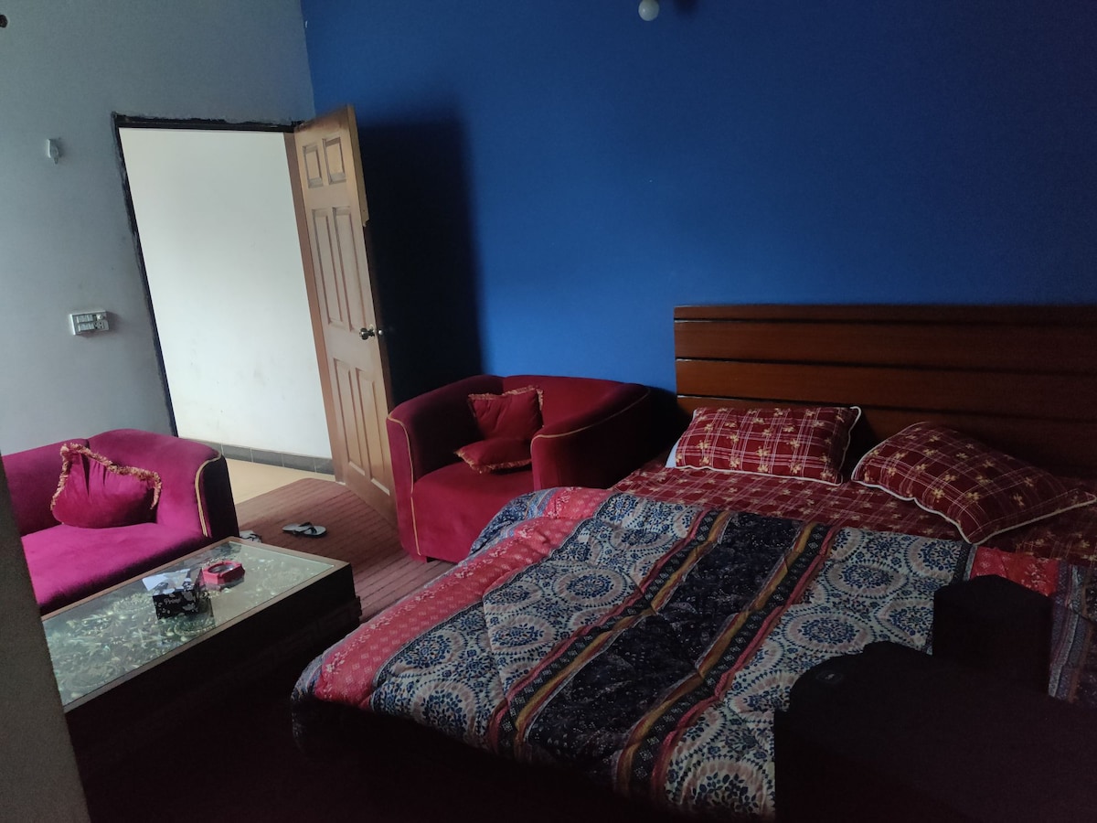 A cozy bedroom features a spacious bed adorned with patterned bedding. Two red armchairs offer additional seating, complemented by a small coffee table. A door leads to another area, and soft lighting creates a relaxing atmosphere.