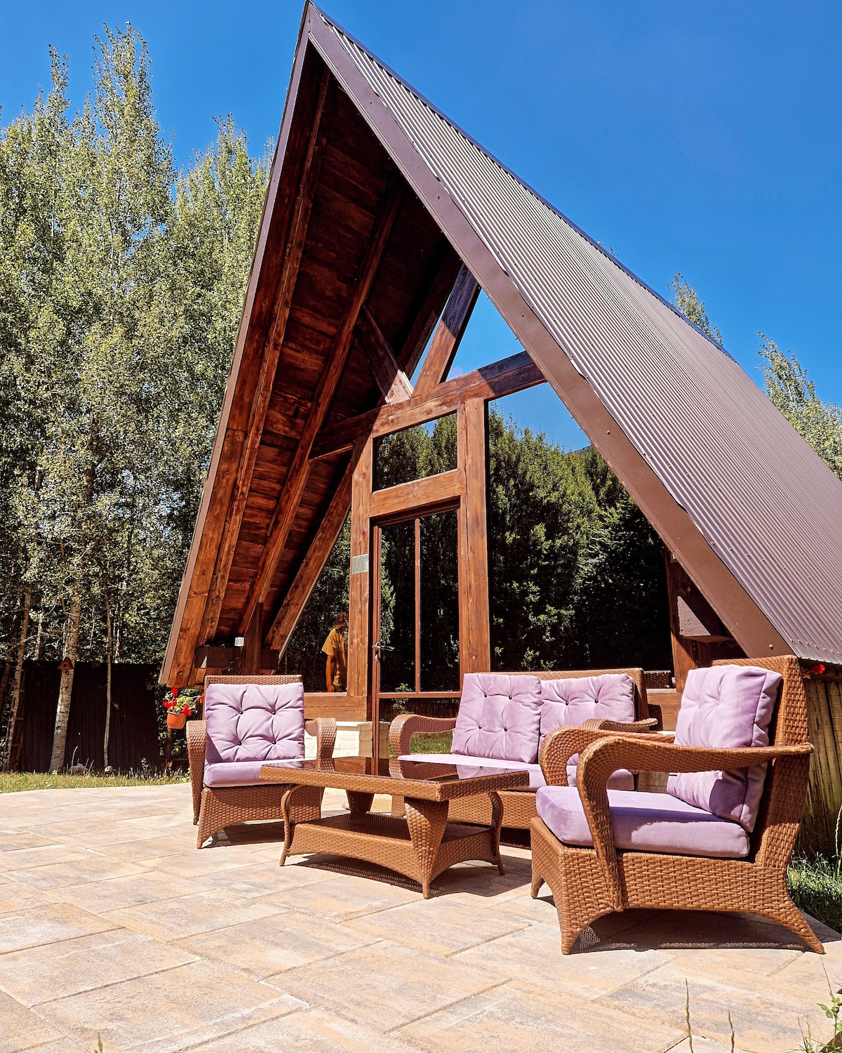 An inviting outdoor seating area is displayed, featuring wicker furniture with soft lavender cushions. The backdrop consists of a cabin with a distinctive A-frame roof, surrounded by lush greenery and clear blue skies, creating a peaceful setting for relaxation.