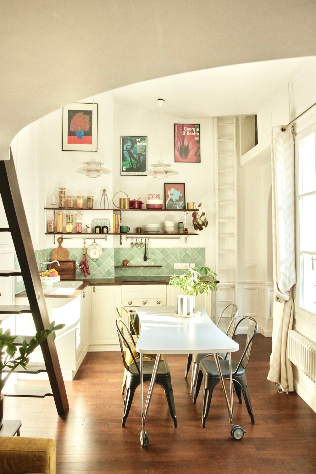 A well-equipped kitchen features a light color palette with a green tile backsplash. A white table is surrounded by four chairs in varying colors. Shelves display jars and decor, while the room is illuminated by natural light streaming through the windows.