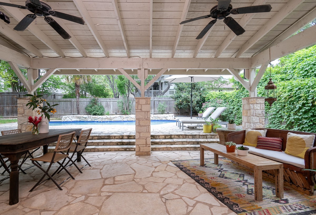 A covered patio area features a dining table surrounded by chairs and a lounge seating arrangement. The textured stone flooring complements the wooden beams of the ceiling. Beyond, an inviting pool area is framed by lush greenery, enhancing the serene outdoor setting.
