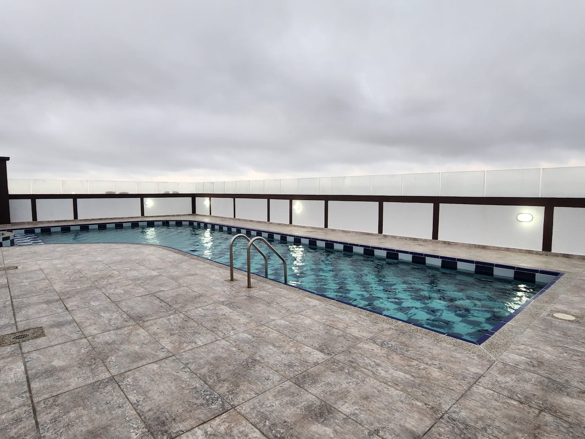 A spacious rooftop pool with clear water is surrounded by a tiled deck. The area is enclosed by a low barrier, providing a sense of privacy. Overcast skies reflect a soft light on the water's surface, creating a tranquil ambiance.