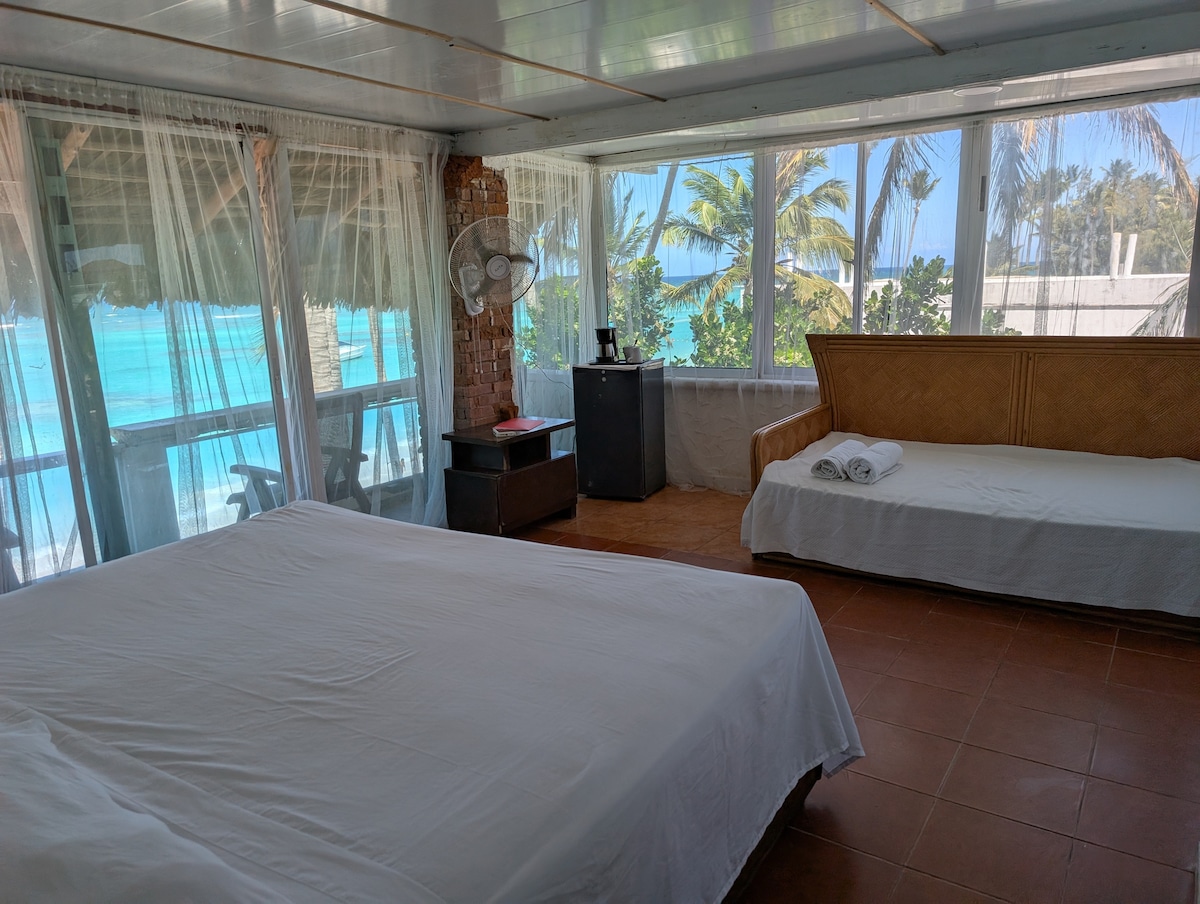 The room features a spacious bed with white bedding and a sofa, surrounded by large windows that provide sweeping views of the beach. Natural light floods the space, highlighting the breezy atmosphere created by palm trees outside.