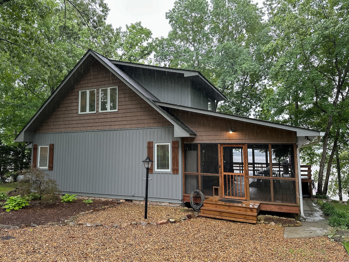 A charming lake house exterior is presented amidst lush greenery. The structure showcases a combination of wooden and grey siding, featuring large windows and an outdoor deck. Gravel pathways meander through landscaped areas, leading to the serene lakeside environment.