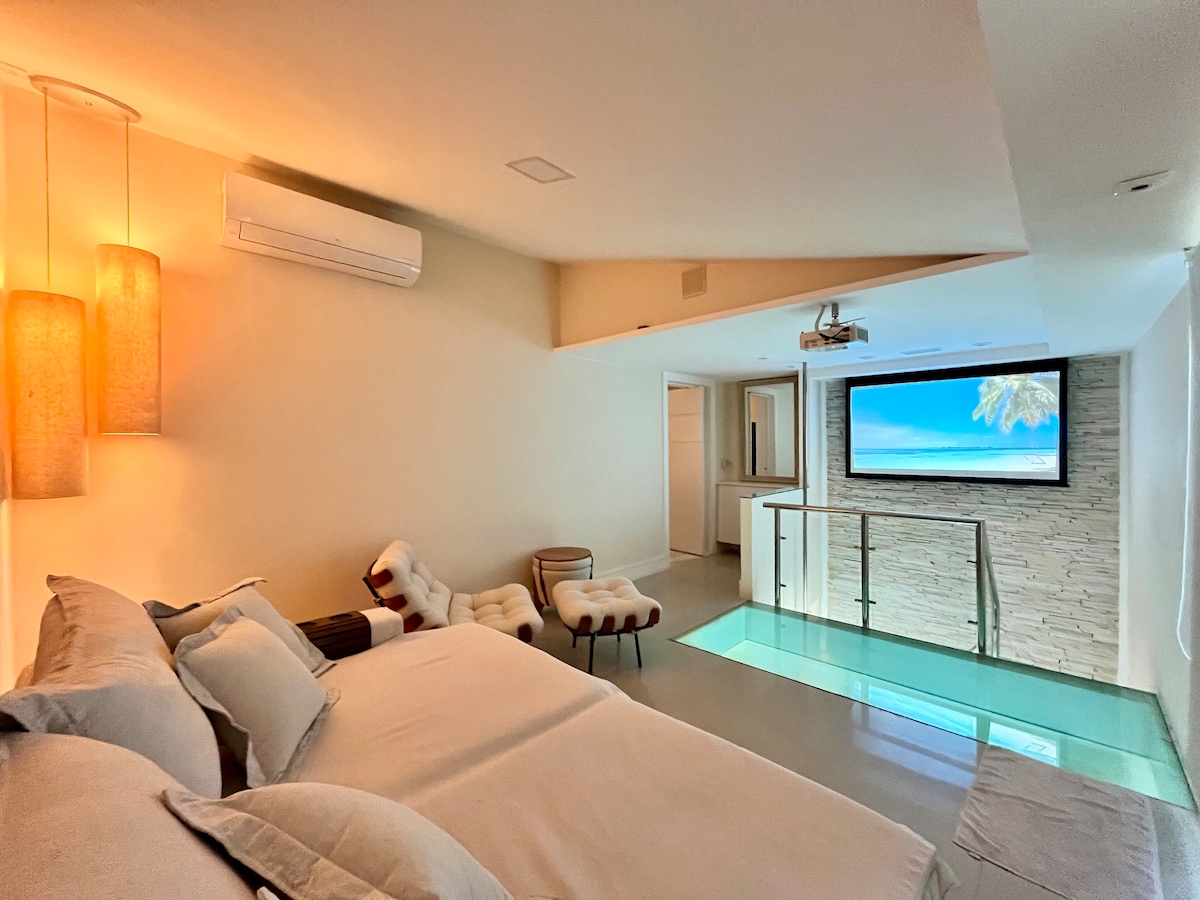 A modern room is shown featuring a spacious bed with neutral-colored bedding. Soft lighting from pendant lamps creates a cozy ambiance. A mounted flat-screen TV and an elegant glass floor section add unique elements, while an air-conditioning unit is visible on the wall.