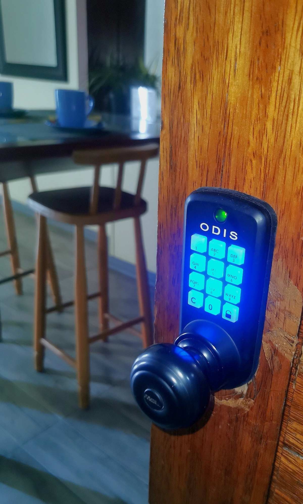 A close-up view of a digital door lock is shown, featuring a blue illuminated keypad with a stylish handle. In the background, wooden bar stools are visible beside a dining table, suggesting a welcoming entryway.
