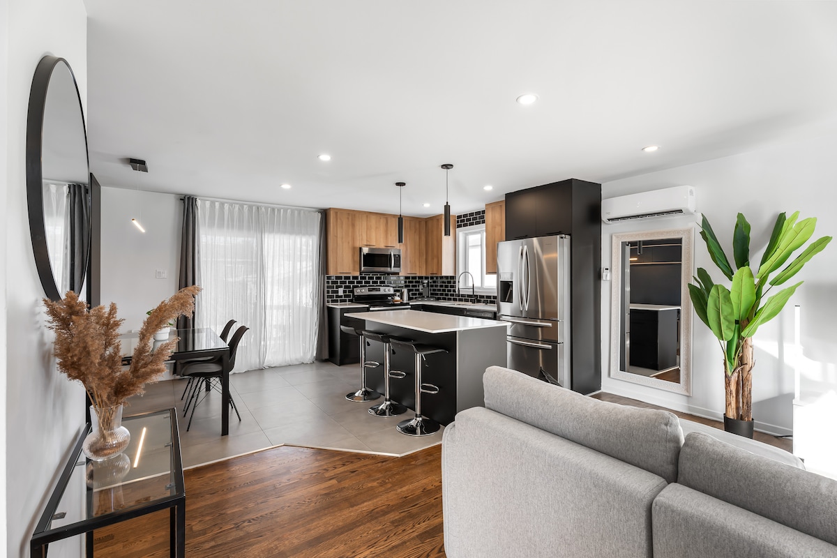 An open-concept living area showcases a modern kitchen equipped with stainless steel appliances and a spacious island. Natural light fills the room through large windows, highlighting a neutral color palette. A cozy sofa and a decorative plant enhance the inviting atmosphere.