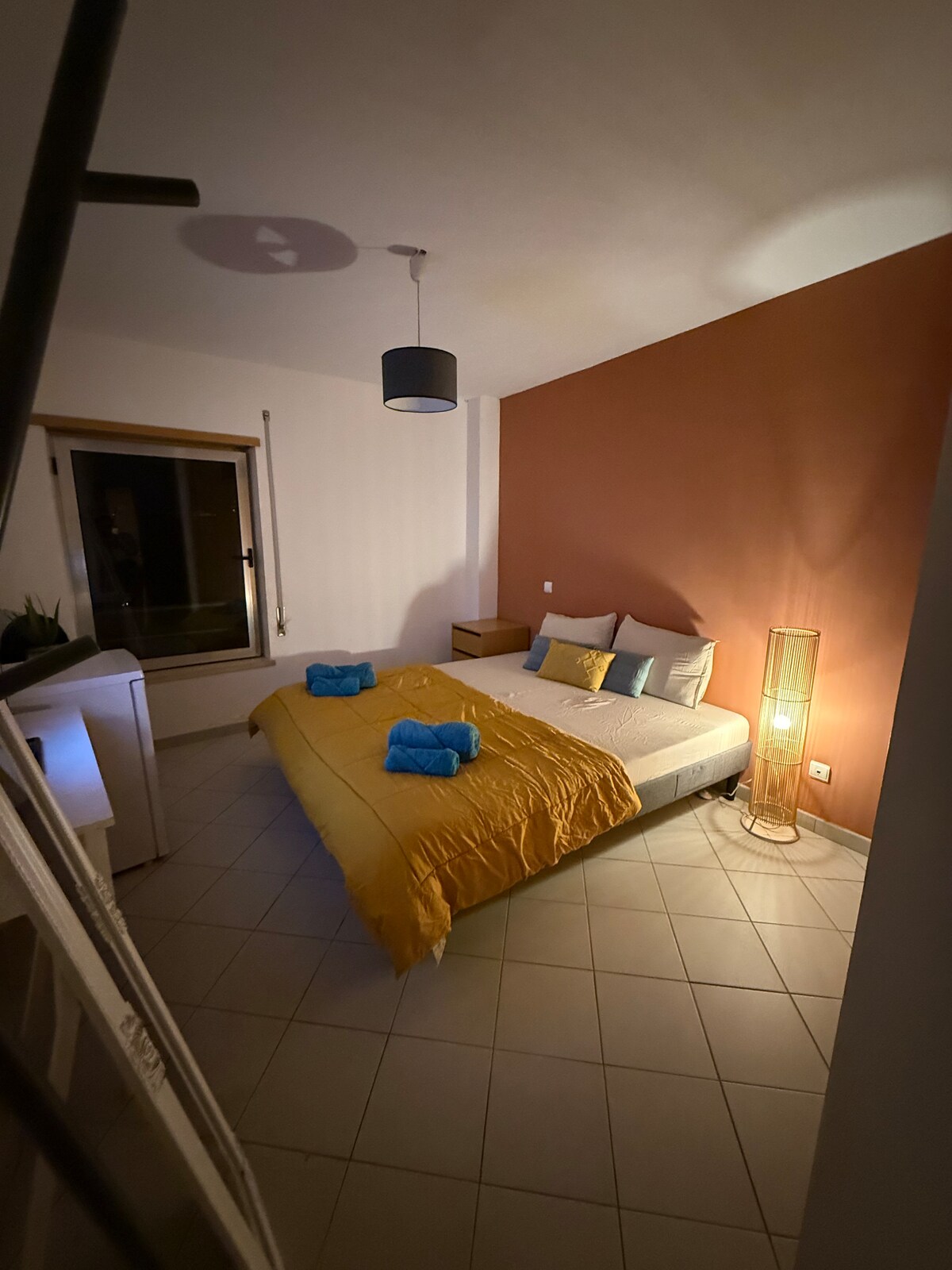 A cozy sleeping area features a spacious bed dressed in a yellow blanket and blue bath towels. A warm-toned accent wall enhances the ambiance. Soft lighting is provided by a floor lamp, and a window offers a glimpse of the outdoor surroundings.