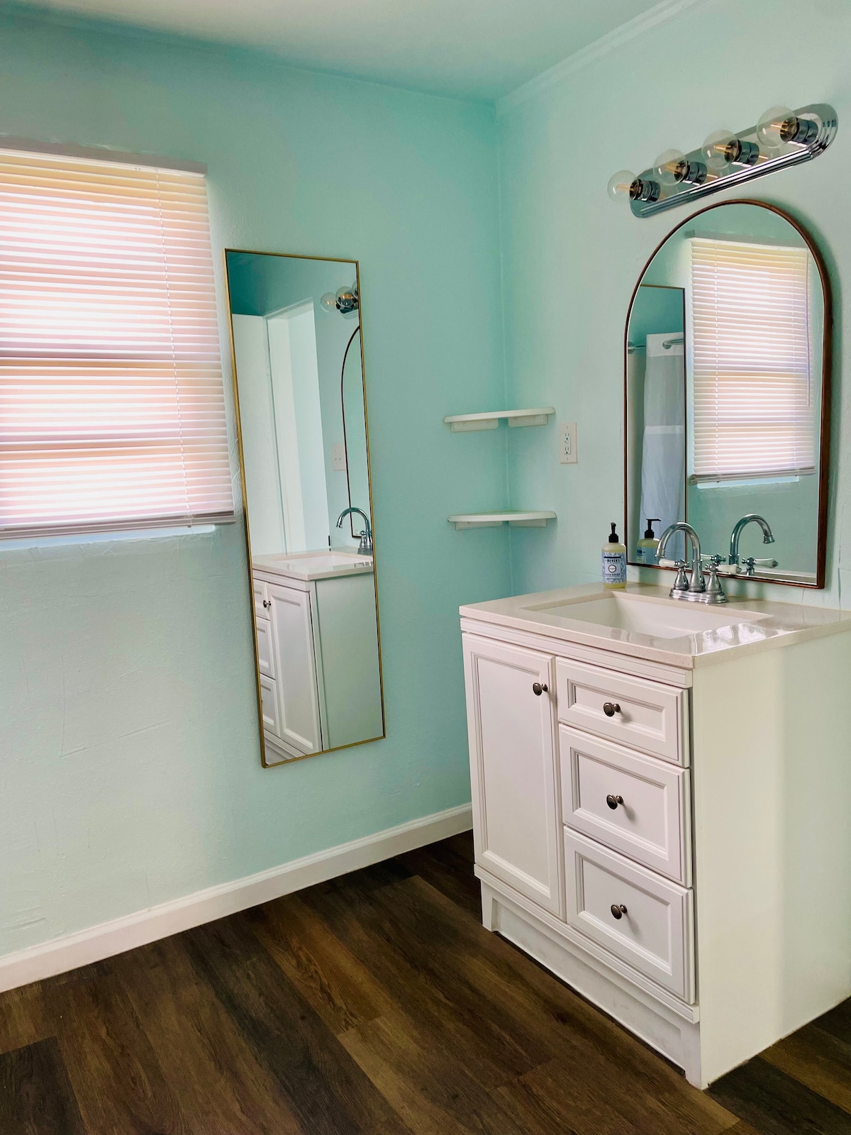 A light blue bathroom features a white vanity with three drawers and a sink, accompanied by a round mirror. Natural light filters through the window, and a shelving unit is mounted beside the mirror, adding functionality to the space.
