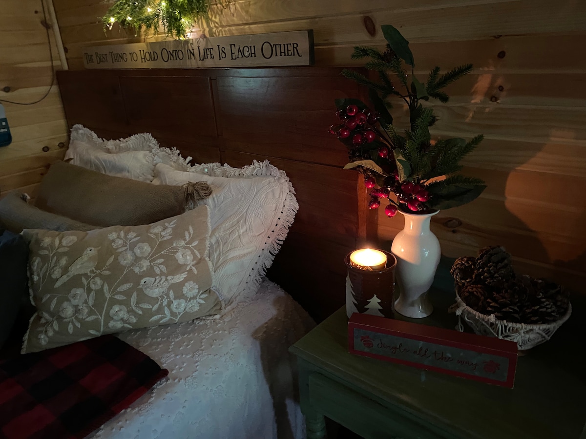 A cozy sleeping area features a textured white coverlet layered on a spacious bed, complemented by decorative pillows. A candle glows softly next to a vase of greenery and festive accents, adding a serene touch to the rustic cabin's interior.