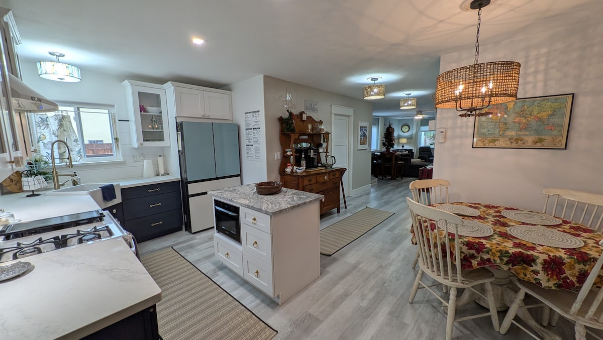 A modern kitchen is showcased, featuring white cabinetry and a large central island with a marble countertop. Stainless steel appliances, including an oven and refrigerator, are visible. A dining area with a round table and light-colored chairs is highlighted, complemented by a decorative chandelier above.