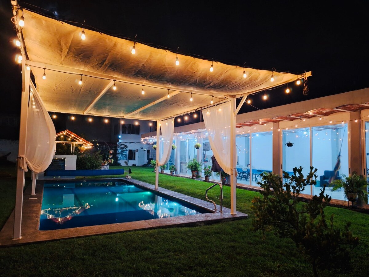A serene nighttime view of a pool area, featuring a partially shaded structure adorned with string lights. The illuminated pool reflects the gentle glow, surrounded by manicured grass. Nearby, transparent walls offer glimpses into the indoor spaces, enhancing the inviting ambiance.