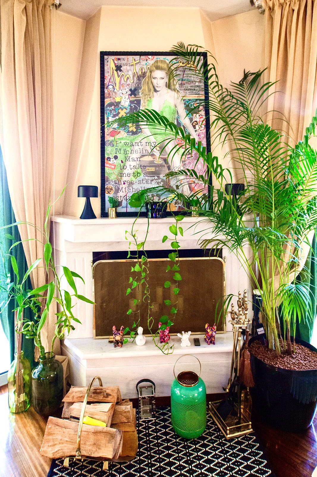 A corner of the living area features a fireplace framed by lush green plants, including tall palm leaves and trailing vines. A textured rug lies beneath, complemented by decorative elements like a green vase and wooden crates.