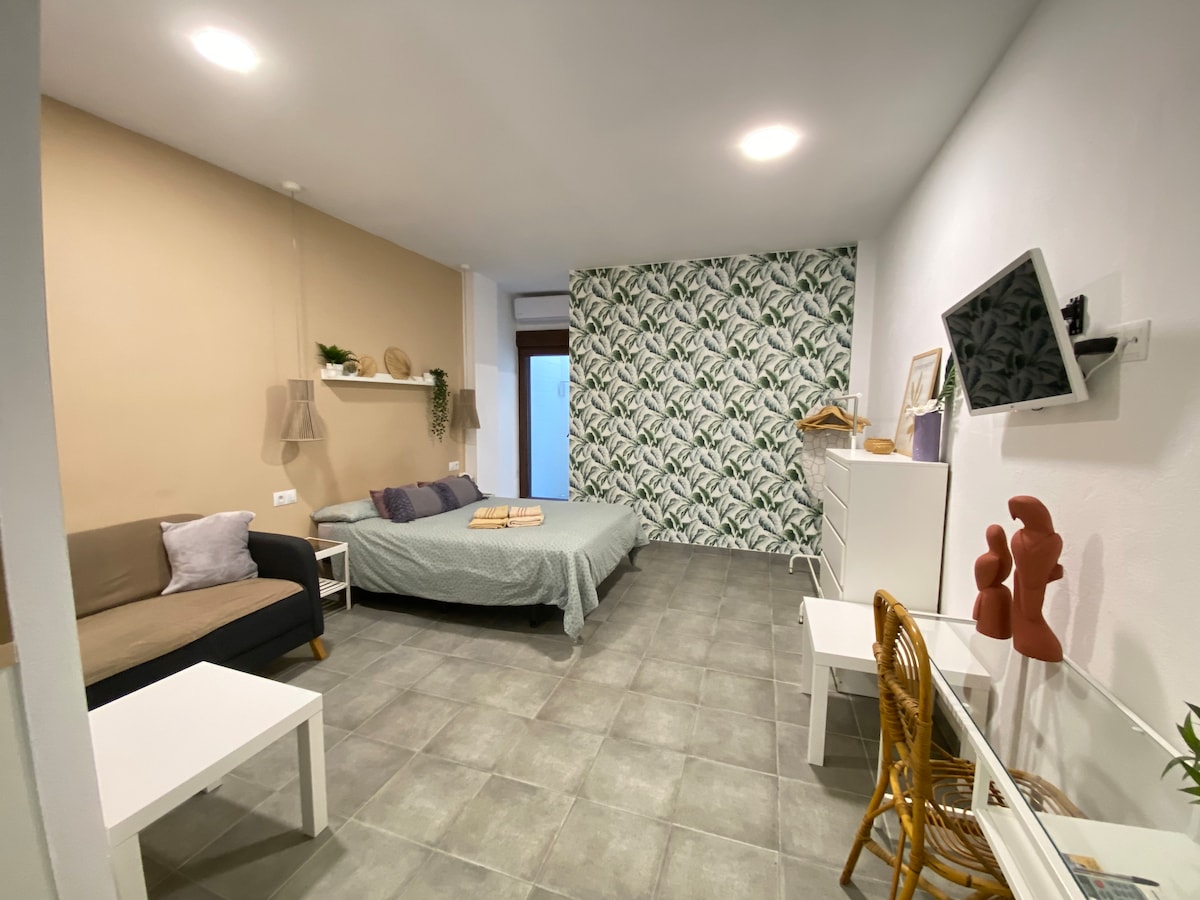 A spacious loft area features a double bed and a sofa, accompanied by a white dresser and a small table with a rattan chair. A vibrant leafy wallpaper creates a fresh backdrop, while natural light enters through the window, enhancing the welcoming atmosphere.