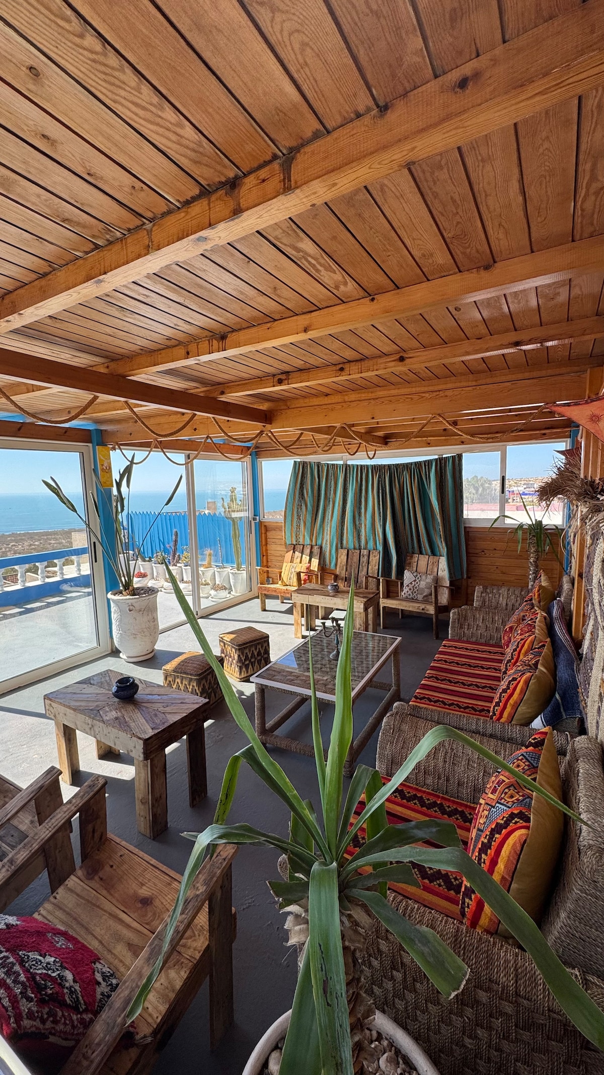 A sunlit sitting area is surrounded by large windows offering a panoramic view of the ocean. Rustic wooden furniture is complemented by colorful textiles on the chairs. Green plants are placed around, adding a touch of nature to the inviting space.