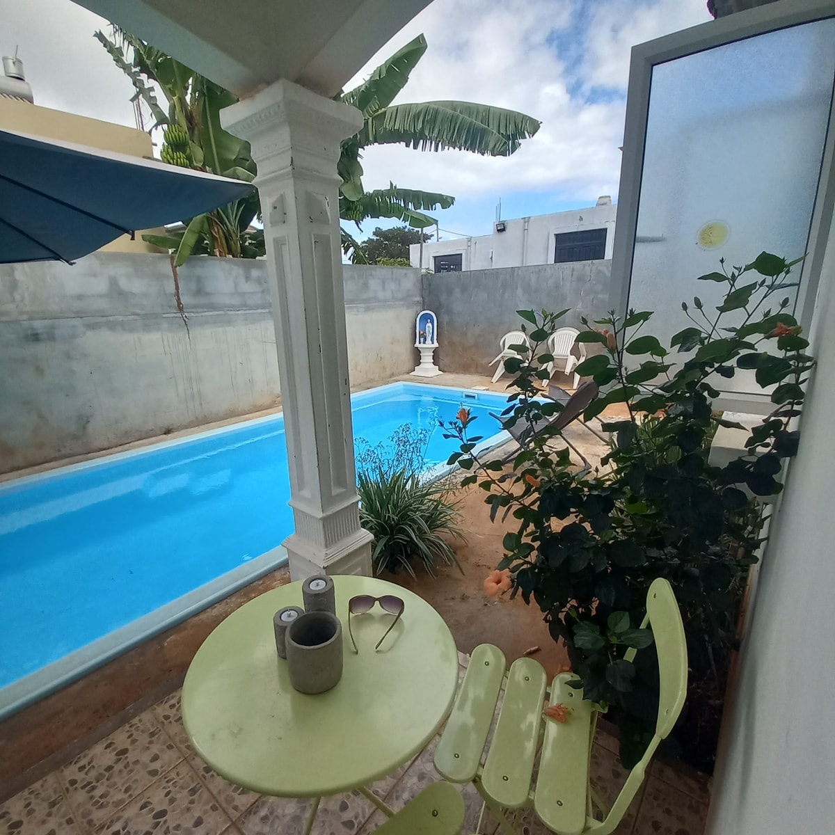 A serene outdoor space features a private pool surrounded by lush greenery. A small green table and chairs are positioned nearby, offering a peaceful seating area. The pool reflects inviting blue water, while the backdrop includes a gently curved wall and tropical plants.