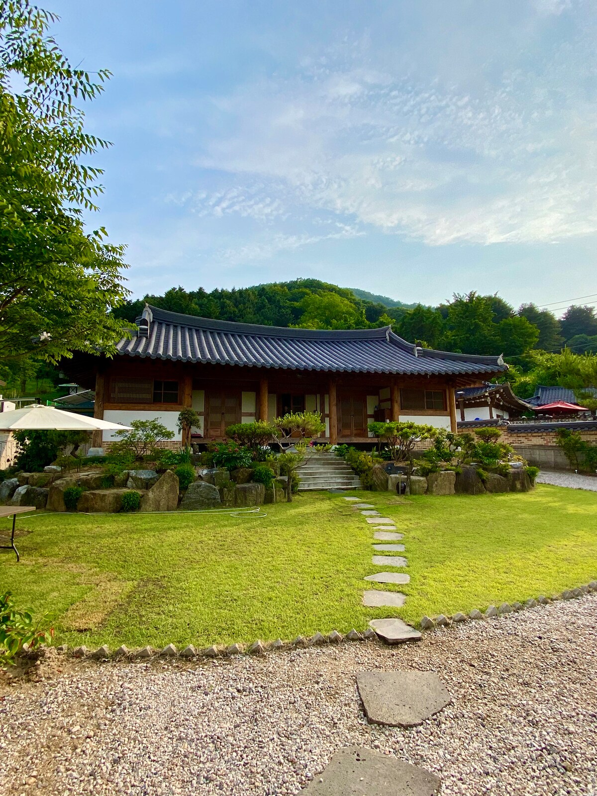 A traditional Korean house is set against a backdrop of lush greenery, featuring a well-maintained garden with stone paths leading to the entrance. The roof displays classic architectural details, while surrounding plants add a serene touch to the outdoor space.