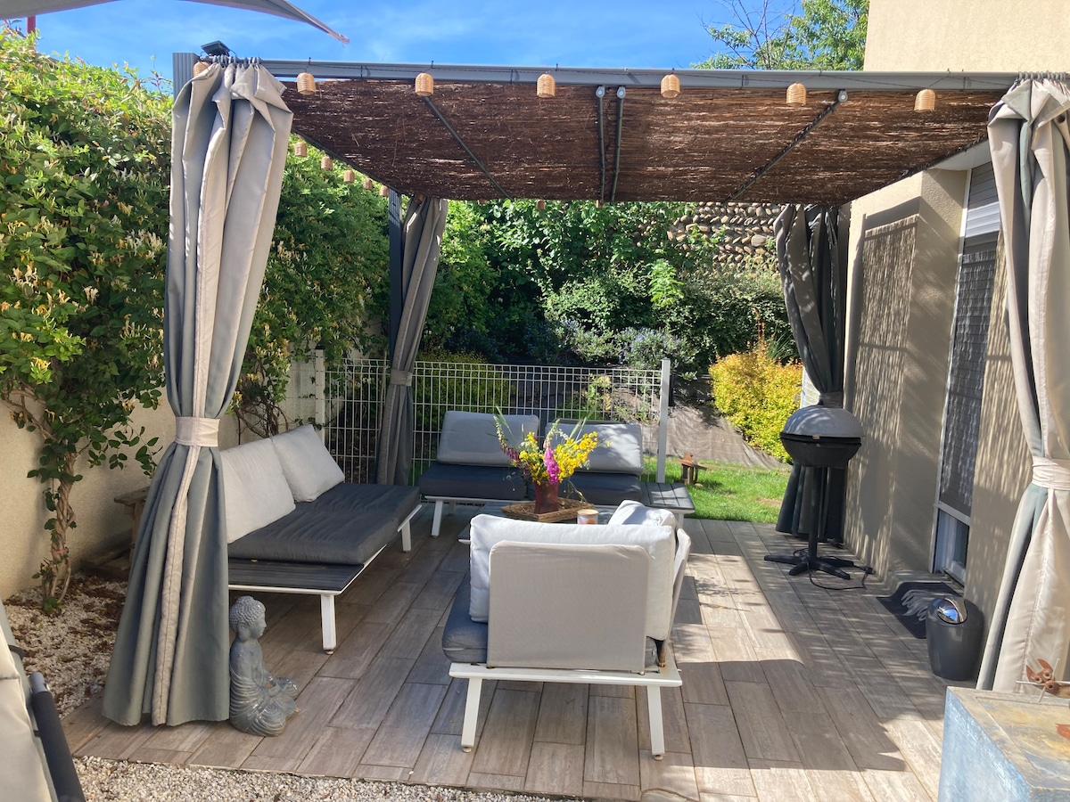 An outdoor seating area is defined by a pergola with draped curtains, creating a shaded space. The area is furnished with a collection of seating, a low table, and a grill. Lush greenery surrounds the space, contributing to a pleasant ambiance.