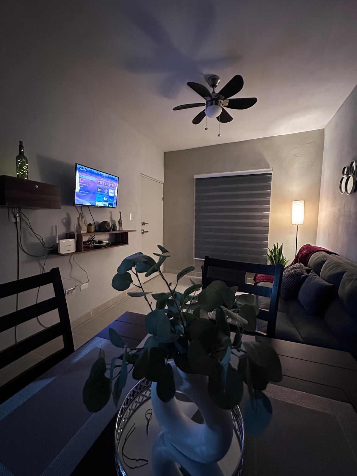 A modern living area is highlighted by a comfortable sofa and a decorative table with a plant centerpiece. A subtle light fixture enhances the ambiance, while a television is mounted on the wall opposite the seating. Neutral colors create a calming environment.