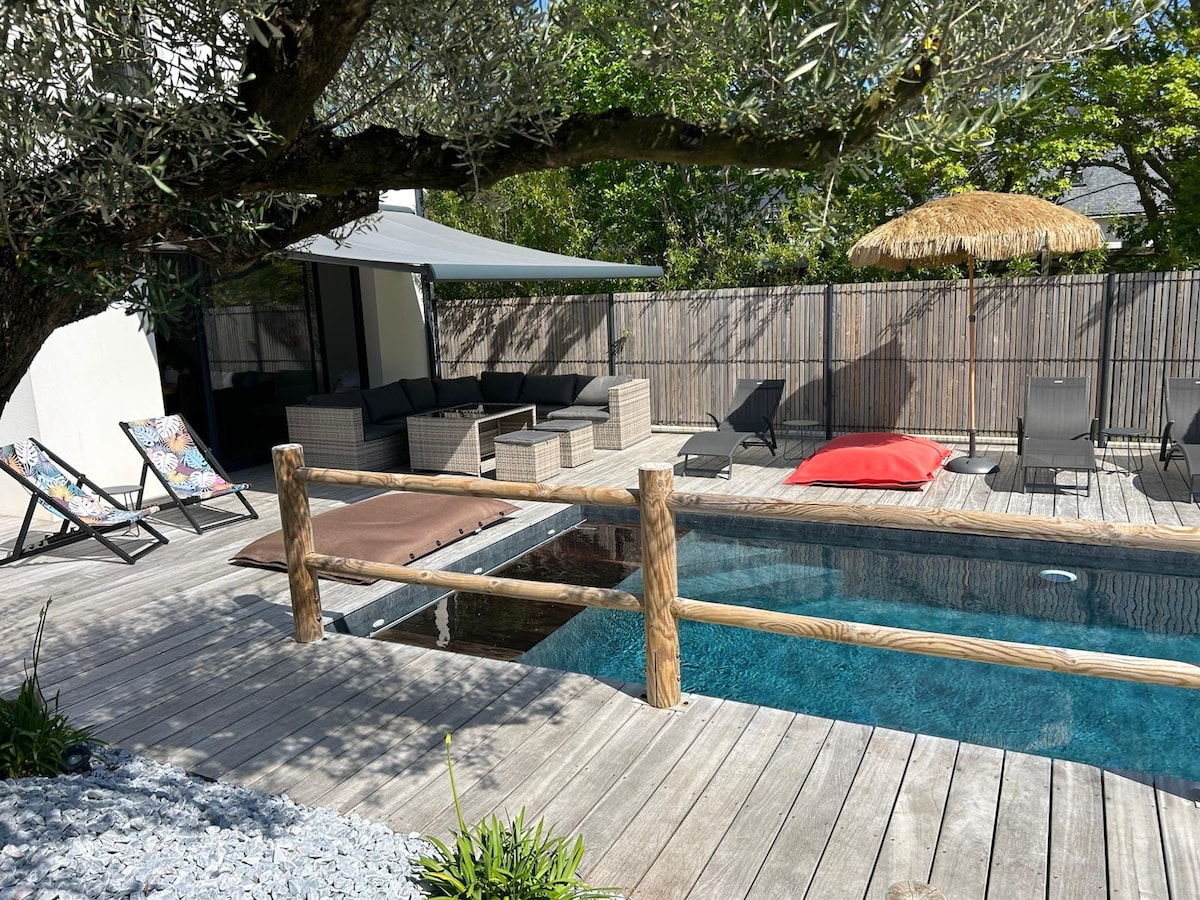 The outdoor area features a saltwater pool surrounded by wooden decking, complemented by lounge chairs and a shaded seating area under a canopy. A bright red sunbed and an umbrella provide additional relaxation spots, with greenery visible in the background.