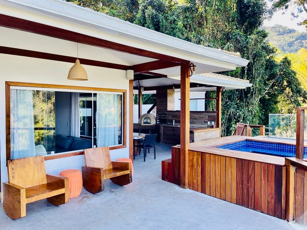 An outdoor area is showcased, featuring a covered space with wooden seating, a small pool, and a barbecue set-up. Large windows allow for natural light, while a scenic backdrop of lush trees adds to the serene environment.