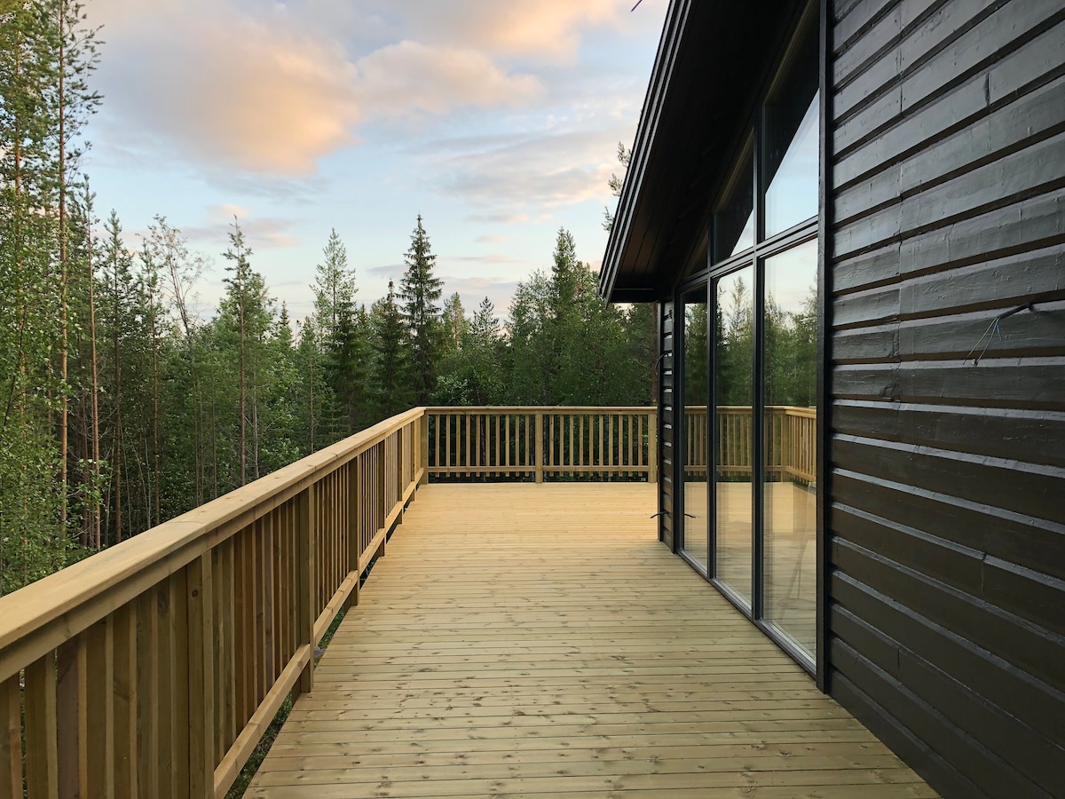 A spacious wooden deck extends from the structure, revealing panoramic views of a wooded area. Large glass windows line the side of the building, allowing natural light to fill the space. The soft colors of the sky create a serene backdrop.