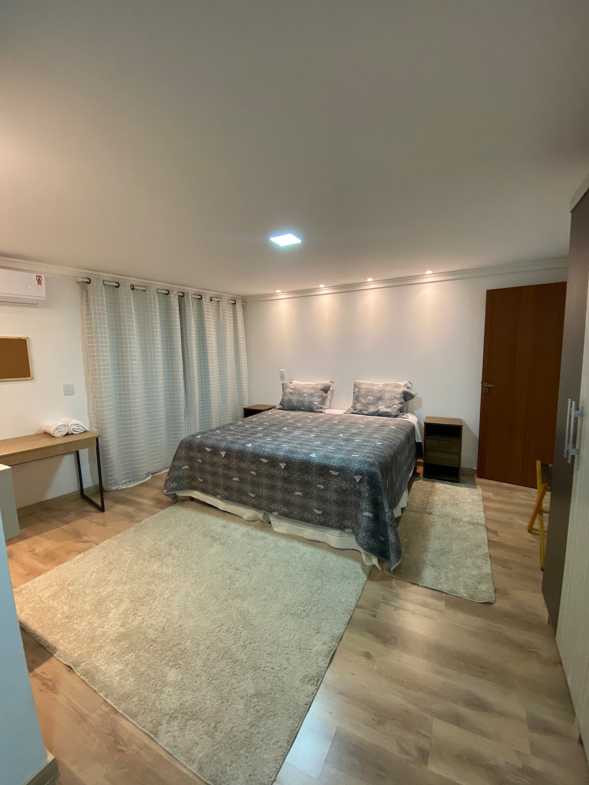 A spacious bedroom features a large bed dressed in neutral linens, flanked by two bedside tables. Soft lighting illuminates the room, complemented by a cozy area rug. A desk with a chair and a wardrobe are present, while sheer curtains allow gentle light to filter in.