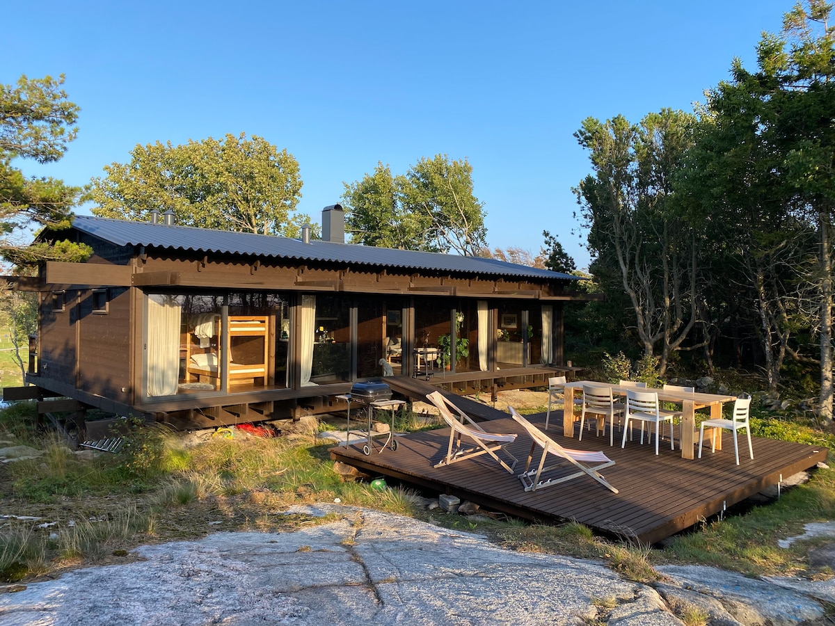 The modern cabin features a wooden deck with multiple lounge chairs and a dining table, set against a natural backdrop of trees and rocky terrain. Large glass doors allow views into the open interior, creating a seamless connection between indoor and outdoor spaces.