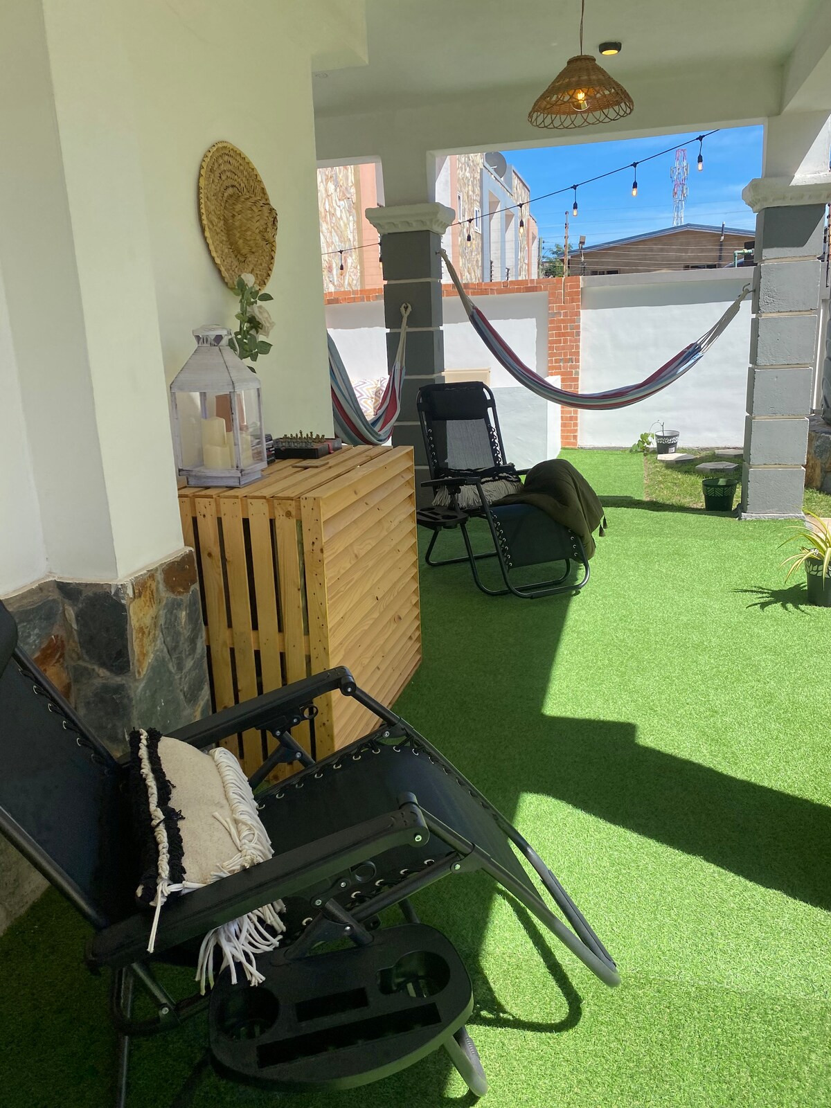 A cozy outdoor space is featured with a green artificial lawn. Two black lounge chairs are positioned beside a wooden storage unit. Above, a hammock is draped between two posts, while warm light fixtures hang from the ceiling, contributing to a relaxing ambiance.