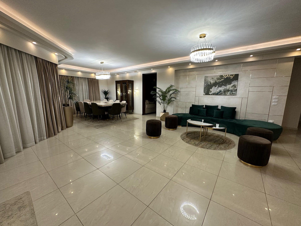 The spacious living area features a modern design with polished tiles and elegant lighting. A green sectional sofa and round ottomans complement the decor. A dining table surrounded by upholstered chairs is visible in the background, and large windows draped with curtains add to the ambiance.