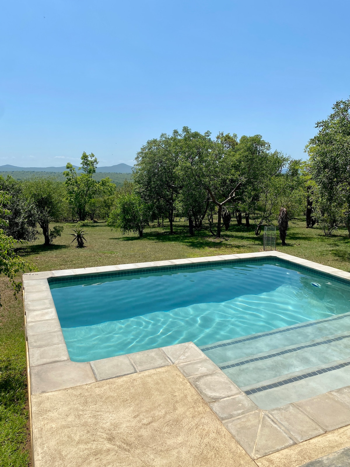 A clear swimming pool reflects sunlight, surrounded by lush green trees and open spaces. The serene landscape showcases distant hills under a clear blue sky, creating a peaceful outdoor setting.