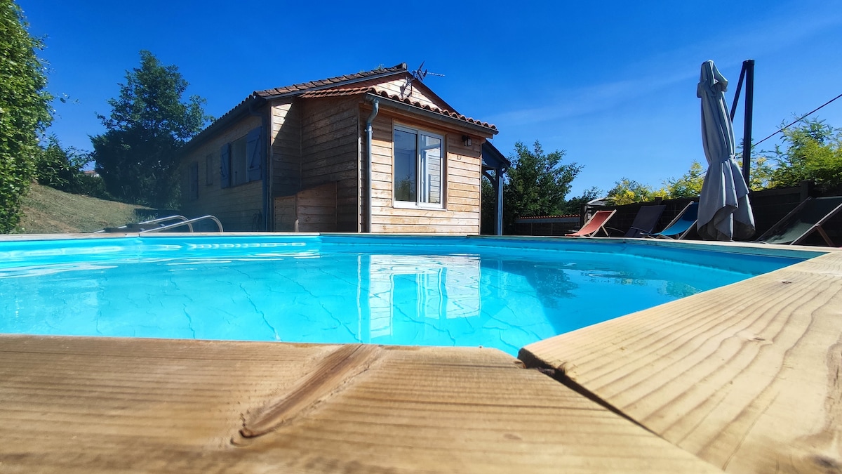 A private semi-buried pool features clear blue water, reflecting the bright sky above. A wooden deck surrounds the pool area, and a sun umbrella is positioned nearby. A wooden maisonette is visible in the background, showcasing multiple windows and a rustic exterior.