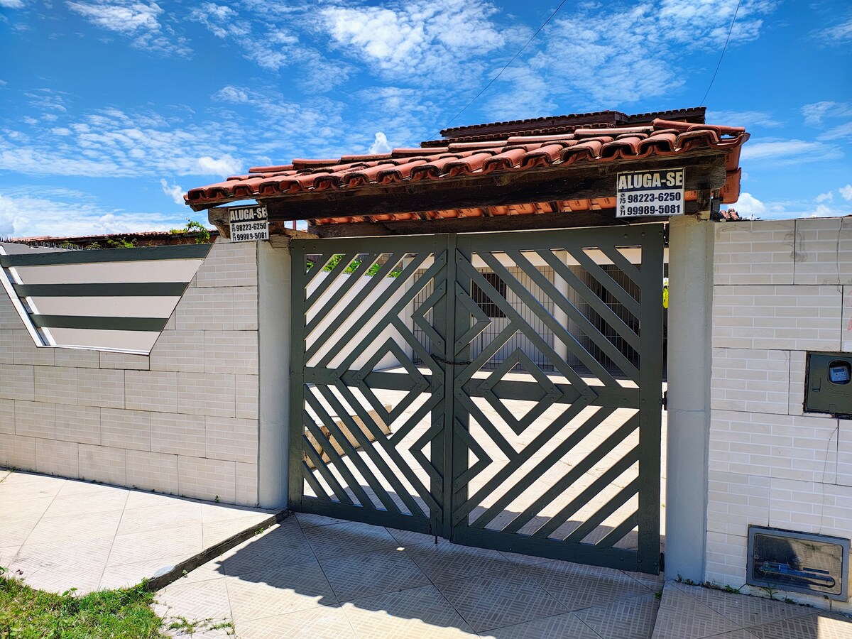 A gated entrance features an intricate design with a wooden canopy above. The gate's green color contrasts with the light-colored brick wall surrounding the property, enhancing the welcoming aspect of the entrance against a clear blue sky.