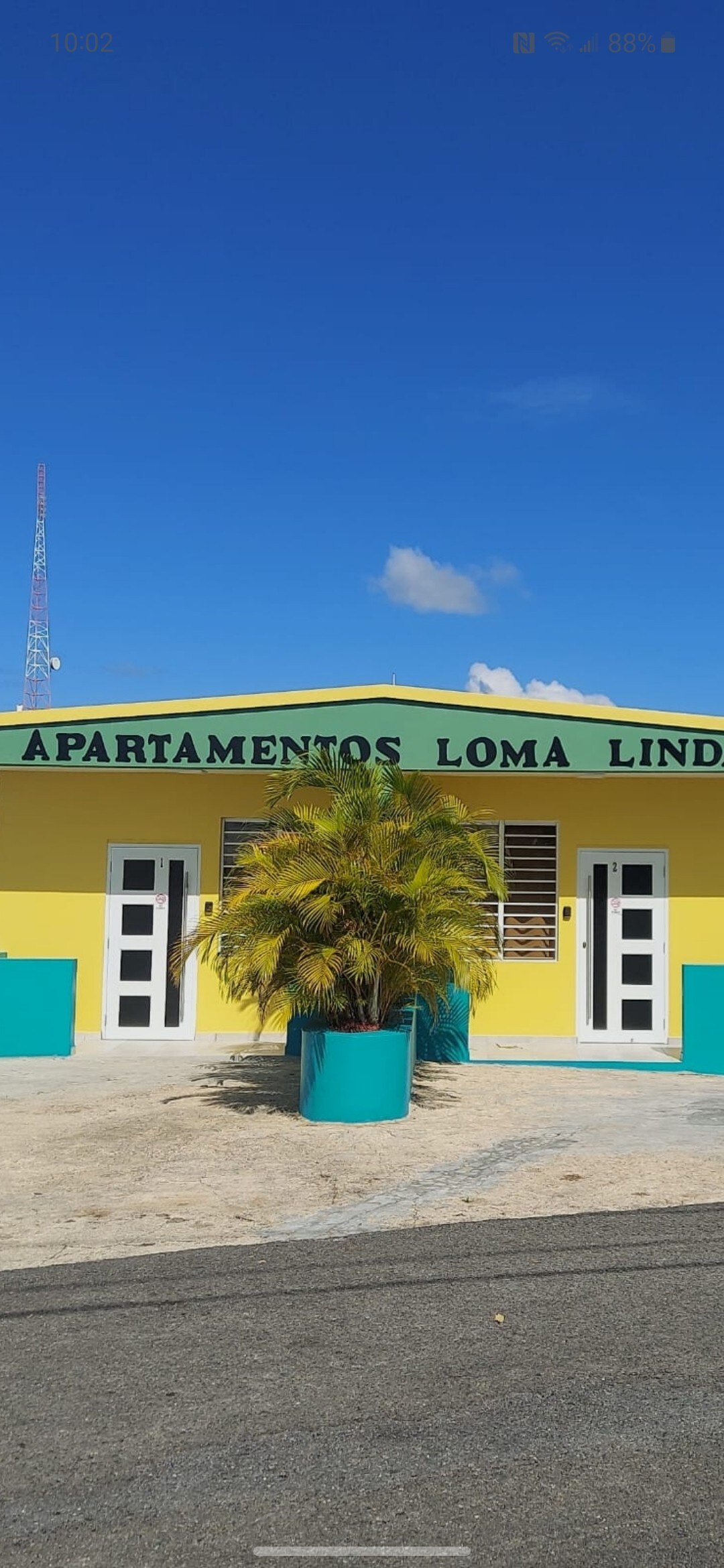 A vibrant yellow building featuring the name 'Apartamentos Loma Linda' prominently displayed above the entrance. Two doors are framed by green planters, while a palm tree adds greenery in front. The clear blue sky complements the colorful facade.