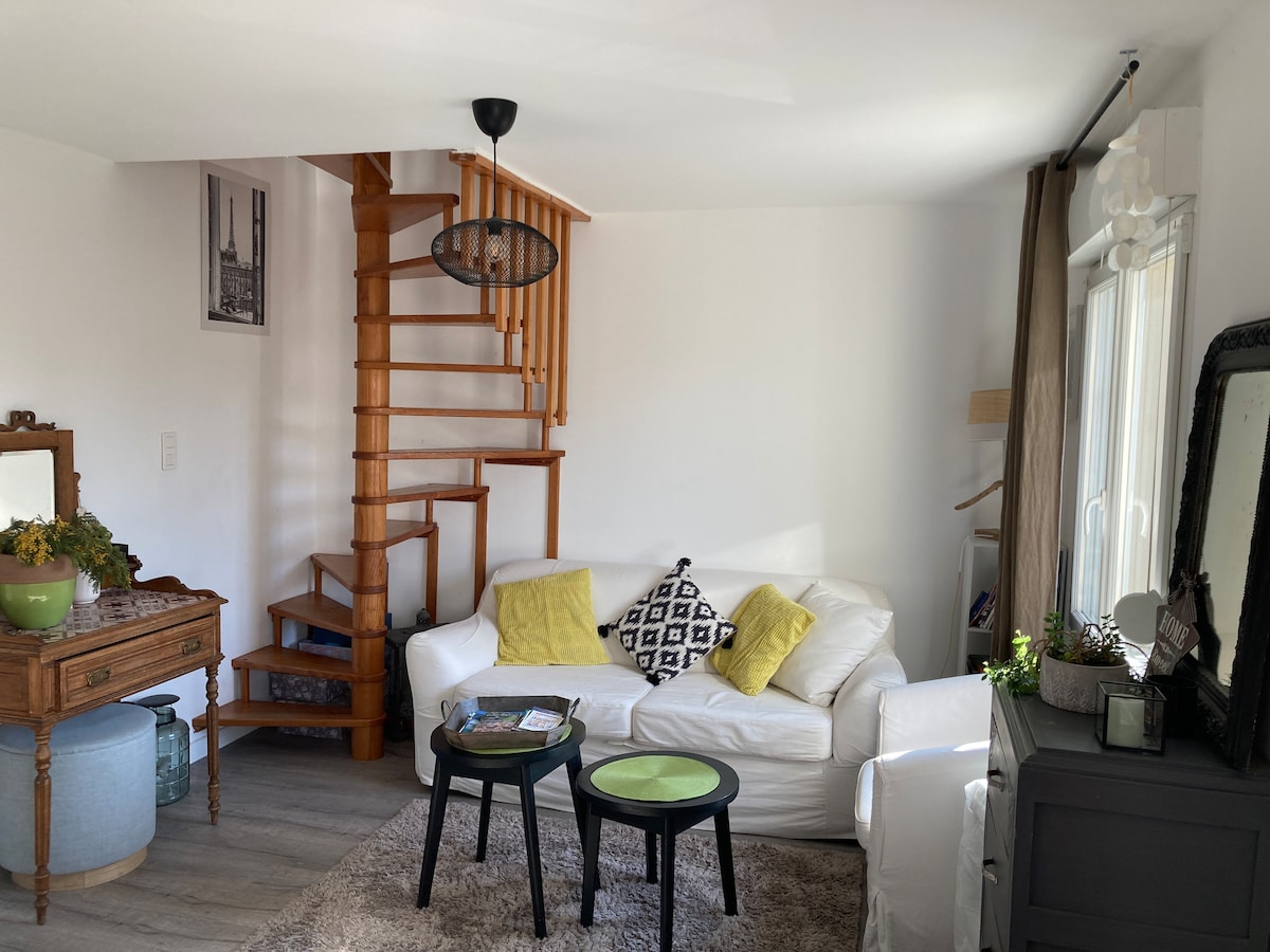 A light-filled living area features a spiral staircase leading to the upper level. A white sofa is complemented by patterned cushions, while a small wooden table and two round tables in green and black are situated in front. A cozy rug covers the floor.