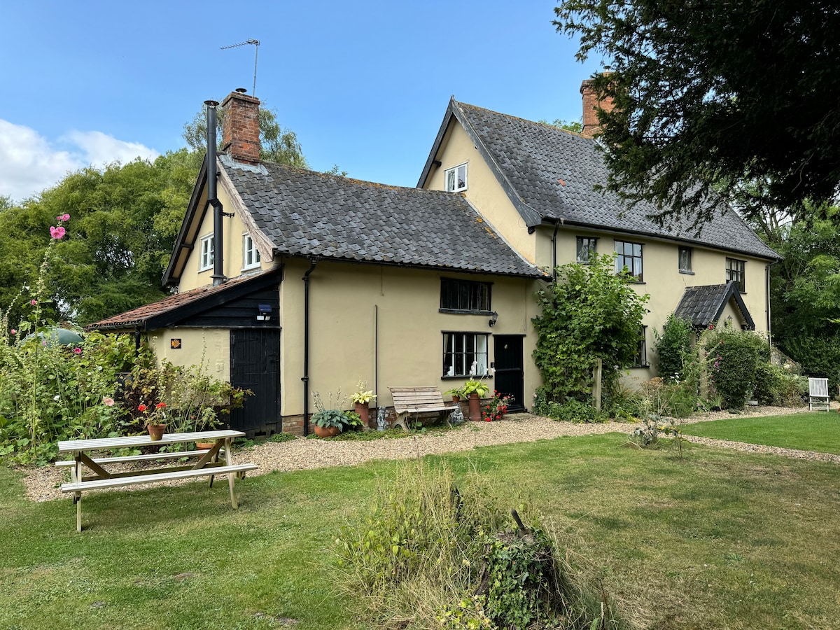The exterior of the Tudor cottage is captured, showcasing a charming, traditional facade with a slate roof and rustic wooden accents. A well-maintained garden features diverse plants and flowers, alongside seating arrangements. Open green space complements the inviting atmosphere of the cottage and surrounding nature.
