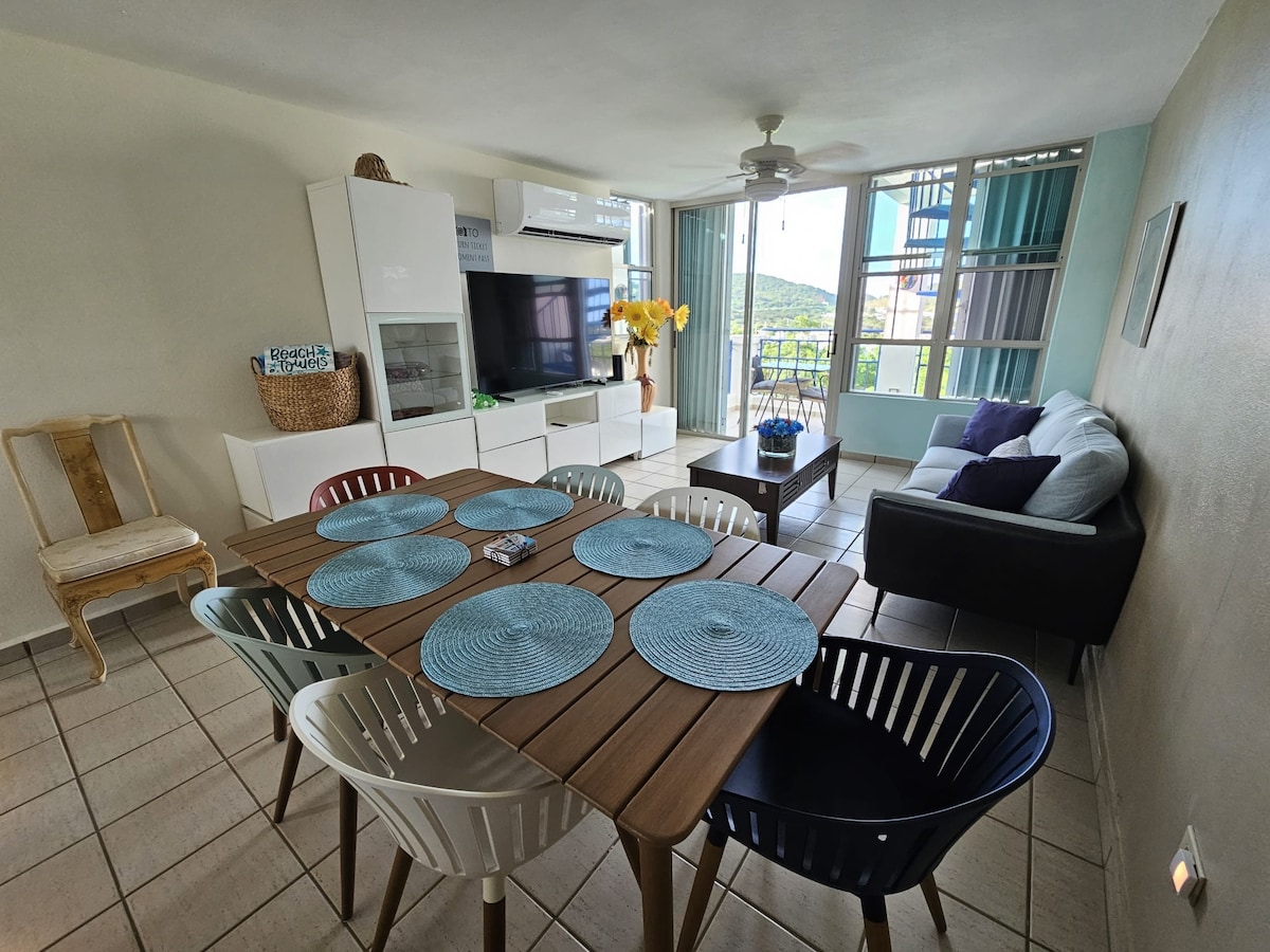 A spacious living and dining area is shown, featuring a wooden table surrounded by colorful chairs. A comfortable sofa and a television are visible, with large windows allowing natural light and views of the outdoors. Neutral-toned walls complement the modern decor.